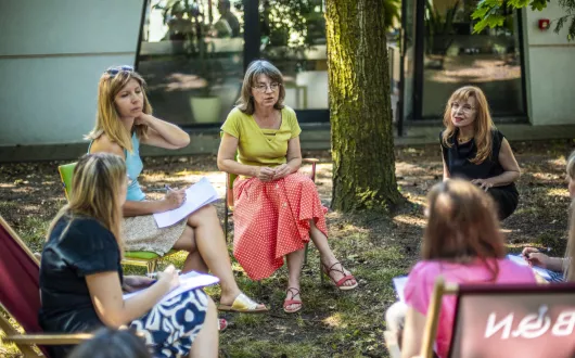 Grupa rozmawiających ze sobą osób siedzących w kręgu na krzesłach i leżakach wśród zieleni.  tyłu widoczny budynek BON. 